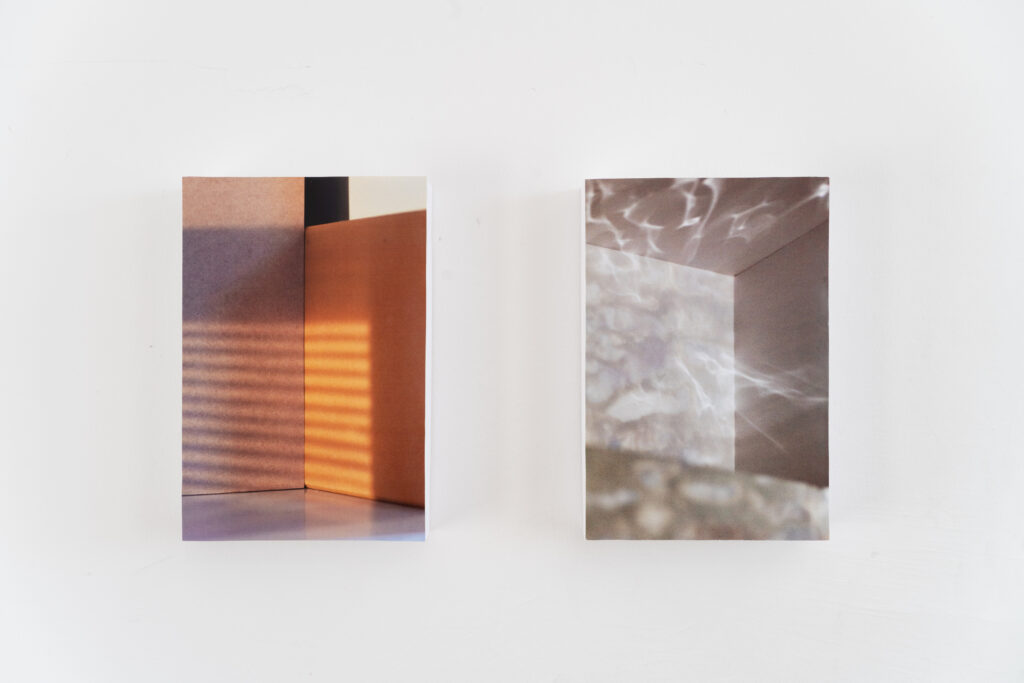 Closeup of two photographs on a wall. The work on the left shows an empty room with lighting coming from outside the image, simulating light shining through blinds. the work on the right also shows an empty room with psychedelic fields of light illuminating the walls, as if reflected from a liquid surface.