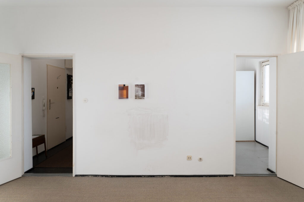 Two photographs on a wall. They hang close to each other, both depicting empty rooms with expressive lighting. Two doors frame the wall. The one on the left leads into a small hallway. The one on the right leads into the kitchen.